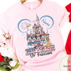 Peut inclure: Un t-shirt rose clair avec un motif Disney. Le design comprend Mickey Mouse, Donald Duck et d'autres personnages en tenue patriotique, avec un ch&acirc;teau et des feux d'artifice. Le texte indique "250 Years Of Freedom" et "Name".
