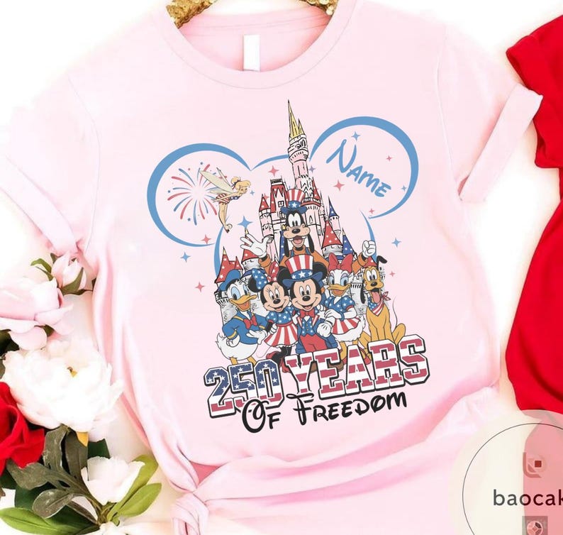 Peut inclure: Un t-shirt rose clair avec un motif Disney. Le design comprend Mickey Mouse, Donald Duck et d'autres personnages en tenue patriotique, avec un ch&acirc;teau et des feux d'artifice. Le texte indique "250 Years Of Freedom" et "Name".