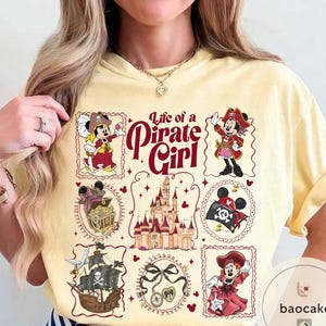 May include: A light yellow t-shirt featuring a "Life of a Pirate Girl" design. The design includes illustrations of Minnie Mouse in pirate attire, a pirate ship, a treasure chest, and a castle. The text is in a burgundy color.