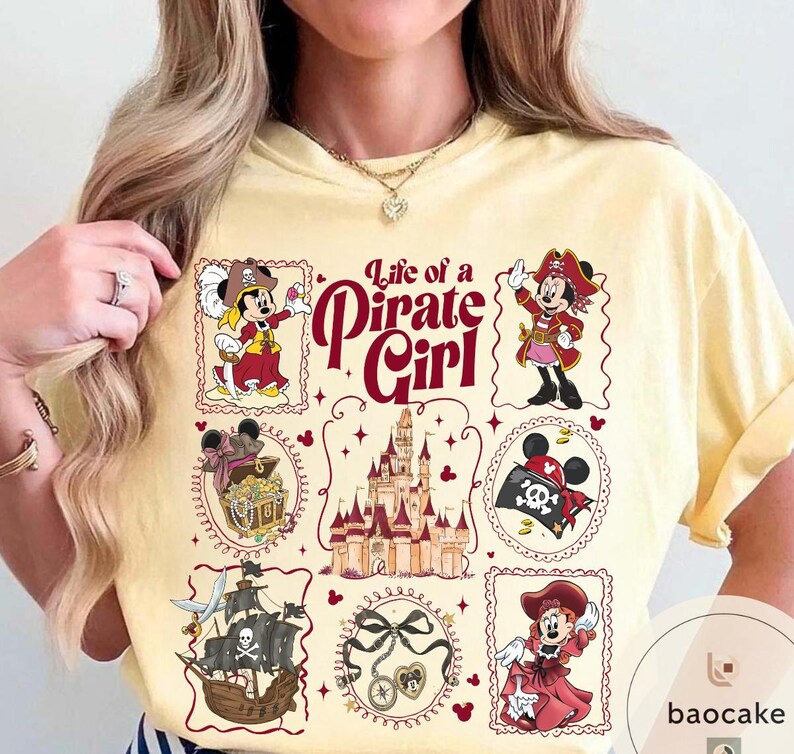 May include: A light yellow t-shirt featuring a "Life of a Pirate Girl" design. The design includes illustrations of Minnie Mouse in pirate attire, a pirate ship, a treasure chest, and a castle. The text is in a burgundy color.