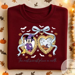 May include: A burgundy crew neck sweatshirt with a Halloween design. The graphic features Minnie Mouse as a witch, pumpkins, bats, and the text "The most scariest place on earth". A light blue ribbon is at the top of the design.