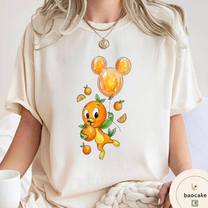 Op de afbeelding: Cr&egrave;mekleurig T-shirt met een cartoon oranje vogel die een sinaasappel en een Mickey Mouse-vormige ballon vasthoudt. De vogel is oranje en geel met groene bladeren. De ballon is oranje. Kleine sinaasappelschijfjes en sinaasappels zijn rond de vogel verspreid.