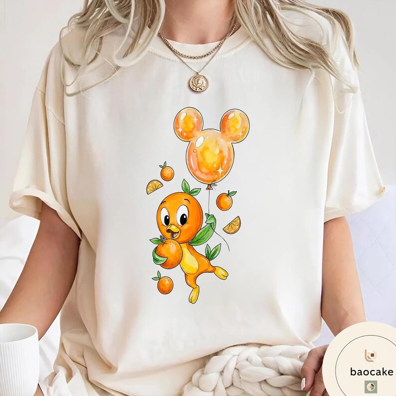 Op de afbeelding: Cr&egrave;mekleurig T-shirt met een cartoon oranje vogel die een sinaasappel en een Mickey Mouse-vormige ballon vasthoudt. De vogel is oranje en geel met groene bladeren. De ballon is oranje. Kleine sinaasappelschijfjes en sinaasappels zijn rond de vogel verspreid.