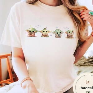 May include: Cream-colored t-shirt featuring a series of four cartoon-style Grogu characters. Each Grogu is depicted in a different pose, with varying colors and details. The design includes small stars and a celestial theme.