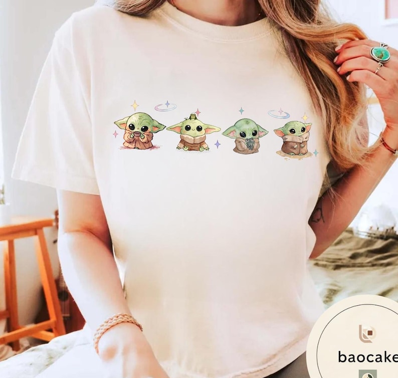 May include: Cream-colored t-shirt featuring a series of four cartoon-style Grogu characters. Each Grogu is depicted in a different pose, with varying colors and details. The design includes small stars and a celestial theme.