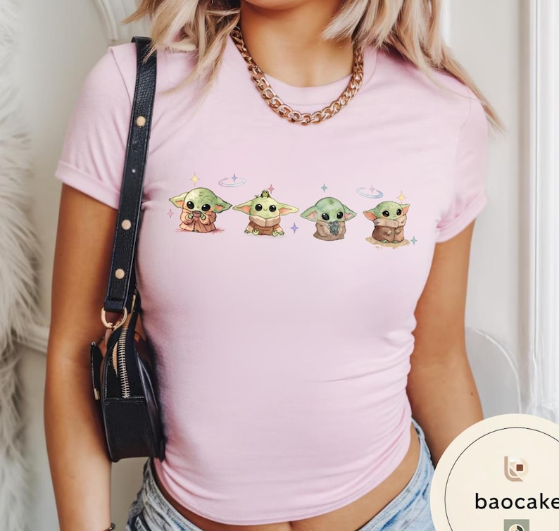 May include: A light pink t-shirt featuring a series of four cartoon Grogu characters. Each Grogu is depicted in a different pose, with a variety of outfits and accessories. The shirt has a crew neck and short sleeves.