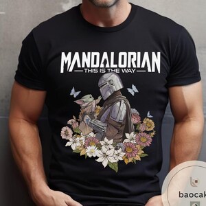 Puede incluir: Camiseta negra con un dise&ntilde;o de The Mandalorian y Grogu rodeados de flores y mariposas. El texto "MANDALORIAN - THIS IS THE WAY" est&aacute; impreso en blanco en la parte superior. El dise&ntilde;o est&aacute; centrado en la camiseta.