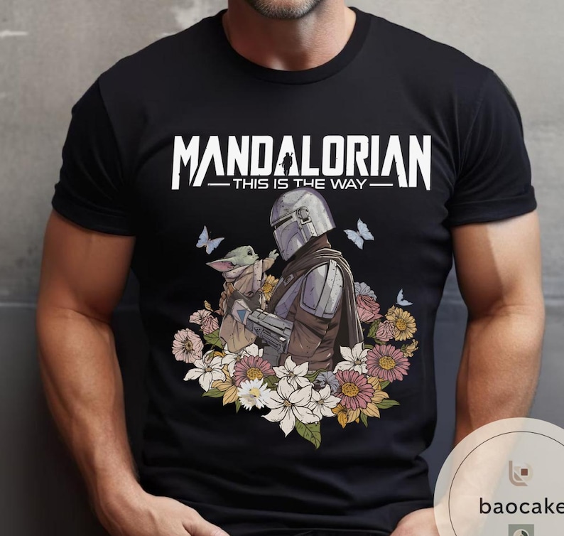 Puede incluir: Camiseta negra con un dise&ntilde;o de The Mandalorian y Grogu rodeados de flores y mariposas. El texto "MANDALORIAN - THIS IS THE WAY" est&aacute; impreso en blanco en la parte superior. El dise&ntilde;o est&aacute; centrado en la camiseta.