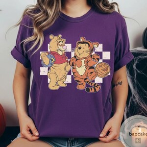 May include: Purple t-shirt featuring a cartoon illustration of two tiger characters. One is dressed in a red shirt and holding a blue bag, while the other is in a tiger costume and holding a jack-o'-lantern. The background is a white and gray checkered pattern.