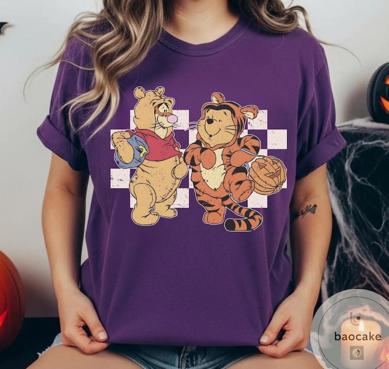 May include: Purple t-shirt featuring a cartoon illustration of two tiger characters. One is dressed in a red shirt and holding a blue bag, while the other is in a tiger costume and holding a jack-o'-lantern. The background is a white and gray checkered pattern.