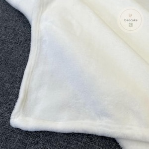 May include: A solid white plush blanket with a soft, fuzzy texture and finished edges. The fabric has a dense, short-pile appearance. A circular logo with the text baocake is visible in the upper right corner of the white fabric surface.