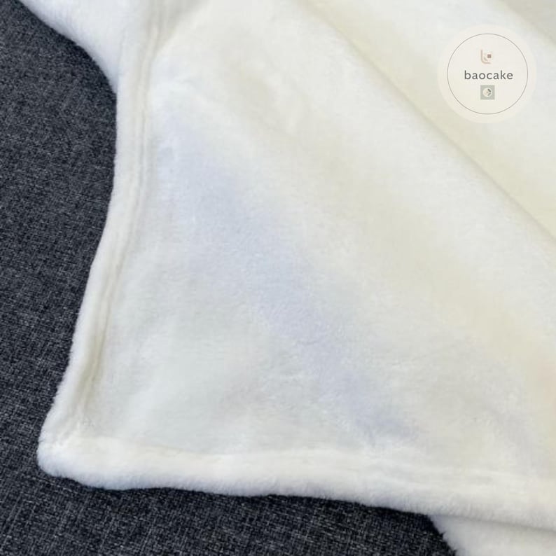 May include: A solid white plush blanket with a soft, fuzzy texture and finished edges. The fabric has a dense, short-pile appearance. A circular logo with the text baocake is visible in the upper right corner of the white fabric surface.