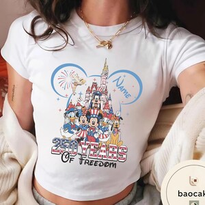 Peut inclure: T-shirt blanc avec un motif color&eacute; de personnages Disney, dont Mickey et Minnie Mouse, devant un ch&acirc;teau. Le design comprend le texte "250 Years of Freedom" et un espace pour un nom, sur le th&egrave;me patriotique.