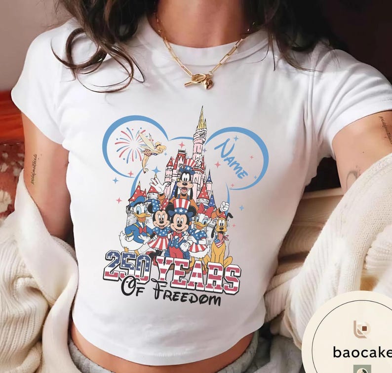 Peut inclure: T-shirt blanc avec un motif color&eacute; de personnages Disney, dont Mickey et Minnie Mouse, devant un ch&acirc;teau. Le design comprend le texte "250 Years of Freedom" et un espace pour un nom, sur le th&egrave;me patriotique.