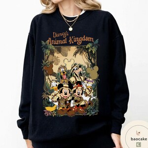 May include: Black sweatshirt featuring a graphic of Disney's Animal Kingdom characters, including Mickey and Minnie Mouse, Donald Duck, and Goofy, in safari attire. The text "Disney's Animal Kingdom" is displayed above the characters in a brown font.