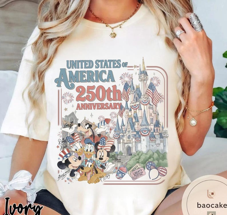 May include: Ivory-colored t-shirt featuring a graphic celebrating the United States of America's 250th Anniversary. The design includes a castle, fireworks, and Disney characters like Mickey and Minnie Mouse, Donald Duck, and Goofy.