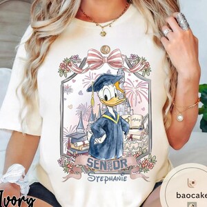 May include: Ivory-colored t-shirt featuring a graduation-themed graphic with Donald Duck in a cap and gown. The design includes a castle, fireworks, and the text "Senior 2023 Stephanie."