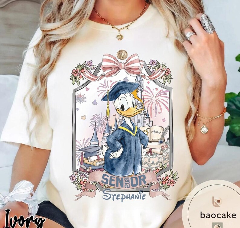 May include: Ivory-colored t-shirt featuring a graduation-themed graphic with Donald Duck in a cap and gown. The design includes a castle, fireworks, and the text "Senior 2023 Stephanie."
