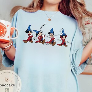 May include: Light blue t-shirt featuring a graphic of Mickey Mouse in wizard attire, with a blue hat and red robe. The design includes a trail of stars. A white mug with an orange sun design is held in the left hand.
