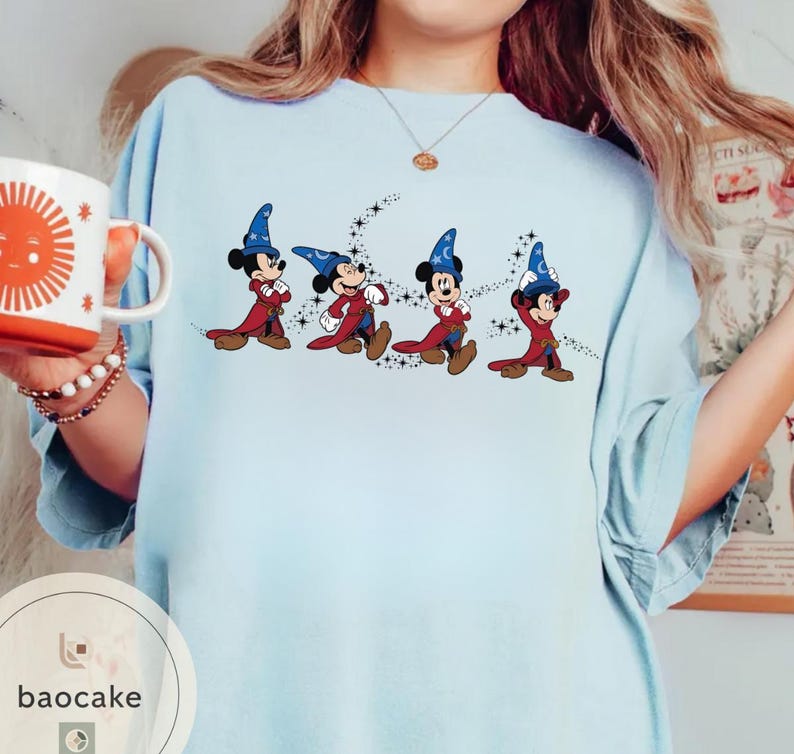 May include: Light blue t-shirt featuring a graphic of Mickey Mouse in wizard attire, with a blue hat and red robe. The design includes a trail of stars. A white mug with an orange sun design is held in the left hand.