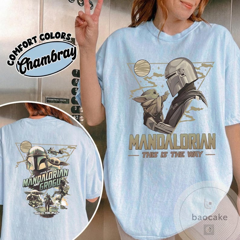 Op de afbeelding: Lichtblauw T-shirt met een afbeelding van The Mandalorian en Grogu, met de tekst "MANDALORIAN - THIS IS THE WAY." De achterkant van het shirt heeft ook een afbeelding van The Mandalorian en Grogu. Het shirt is gelabeld "Comfort Colors Chambray."