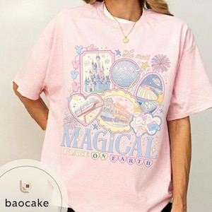 May include: A pale pink t-shirt featuring a vibrant graphic. The design incorporates images of a castle, a globe, a monorail, and other theme park attractions. The words "MAGICAL PLACE ON EARTH" are written in light blue.