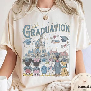May include: Cream-colored t-shirt featuring the word "Graduation" in blue script above a cartoon illustration. The illustration includes a castle, graduation caps, diplomas, and Disney characters Mickey, Minnie, Donald, Daisy, Goofy, and Pluto.