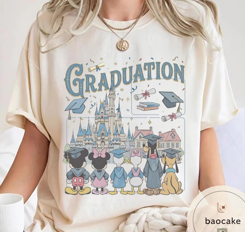 May include: Cream-colored t-shirt featuring the word "Graduation" in blue script above a cartoon illustration. The illustration includes a castle, graduation caps, diplomas, and Disney characters Mickey, Minnie, Donald, Daisy, Goofy, and Pluto.