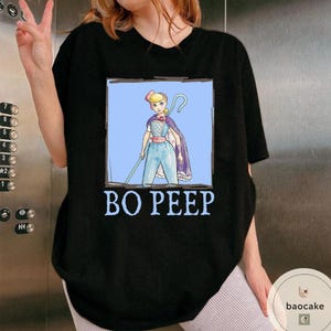 K&ouml;nnte beinhalten: Ein schwarzes Kurzarm-T-Shirt mit einer rechteckigen Grafik einer Zeichentrickfigur mit blonden Haaren, die ein blaues Outfit und einen violetten Umhang tr&auml;gt und einen Hirtenstab h&auml;lt. Der Text BO PEEP ist in Hellblau unter der Grafik gedruckt.
