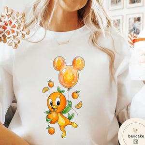 Op de afbeelding: Witte sweatshirt met een cartoon sinaasappel karakter met een sinaasappel en een Mickey Mouse-vormige ballon. Het ontwerp bevat sinaasappelschijfjes en kleine sinaasappels. Het karakter is oranje en geel met groene bladeren.