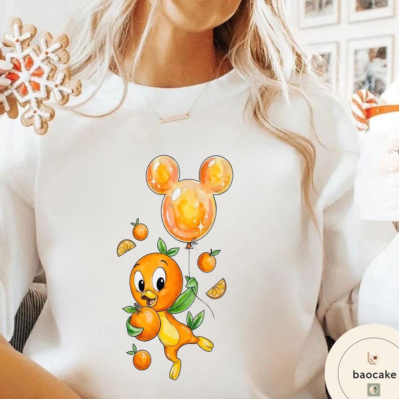 Op de afbeelding: Witte sweatshirt met een cartoon sinaasappel karakter met een sinaasappel en een Mickey Mouse-vormige ballon. Het ontwerp bevat sinaasappelschijfjes en kleine sinaasappels. Het karakter is oranje en geel met groene bladeren.