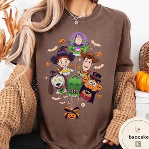 Pode incluir: Uma t-shirt castanha com uma impress&atilde;o gr&aacute;fica de personagens de Toy Story vestidos com fatos de Halloween, incluindo Buzz Lightyear, Woody, Jessie, Rex, Slinky Dog e Mr. Potato Head, atados como bal&otilde;es com uma ab&oacute;bora na base.
