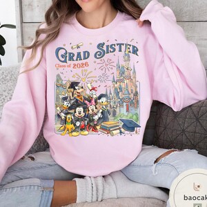 May include: Pale pink sweatshirt featuring the words "Grad Sister Class of 2026" and a Disney-themed graduation graphic. The design includes characters and a castle. The sweatshirt has a crew neck and long sleeves.