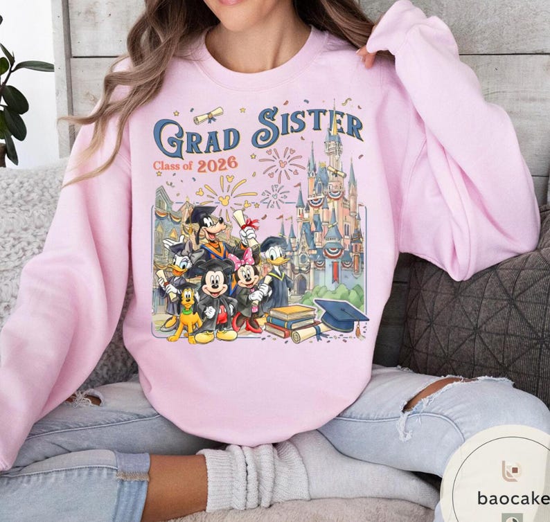 May include: Pale pink sweatshirt featuring the words "Grad Sister Class of 2026" and a Disney-themed graduation graphic. The design includes characters and a castle. The sweatshirt has a crew neck and long sleeves.