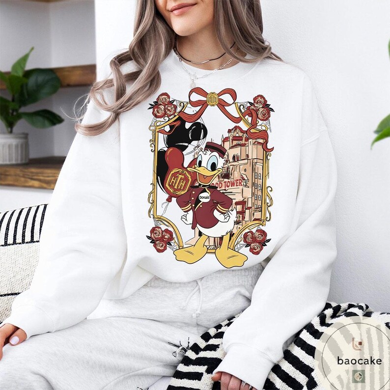 May include: White sweatshirt featuring a framed graphic of Donald Duck in a maroon uniform holding a balloon. The graphic includes the Hollywood Tower Hotel and red roses. The sweatshirt has long sleeves and a crew neck.