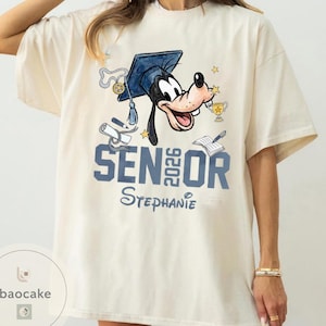 May include: Cream-colored t-shirt featuring a cartoon of Goofy wearing a graduation cap. The shirt has the text "SENIOR 2026 Stephanie" in blue. Graduation-themed graphics, including a diploma and trophy, are also present.