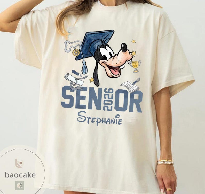 May include: Cream-colored t-shirt featuring a cartoon of Goofy wearing a graduation cap. The shirt has the text "SENIOR 2026 Stephanie" in blue. Graduation-themed graphics, including a diploma and trophy, are also present.