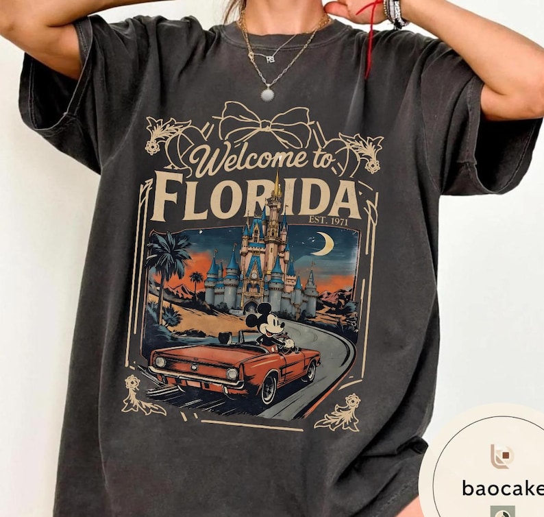 May include: Dark gray t-shirt with a graphic that says "Welcome to Florida" and features a vintage-style illustration of a car with Mickey Mouse driving past a castle. The shirt has a relaxed fit.