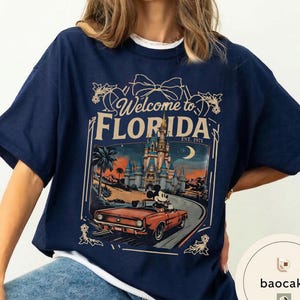 May include: Navy blue t-shirt with a graphic featuring Mickey Mouse driving a red convertible in front of a castle. The text "Welcome to Florida" and "Est. 1971" are also displayed on the shirt.