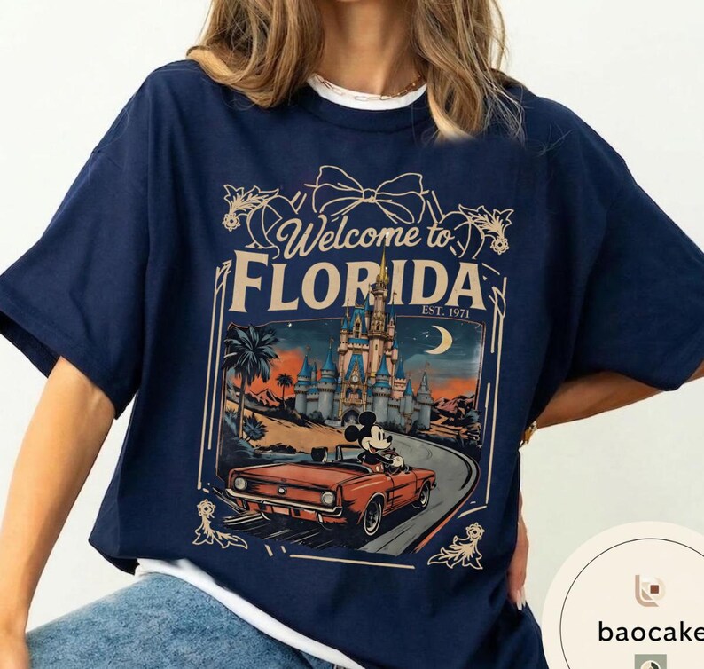 May include: Navy blue t-shirt with a graphic featuring Mickey Mouse driving a red convertible in front of a castle. The text "Welcome to Florida" and "Est. 1971" are also displayed on the shirt.