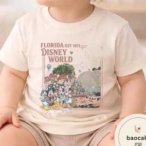 May include: A cream-colored short-sleeve t-shirt featuring a graphic print of Disney World landmarks and characters. The text reads Florida Est 1971 Disney World The Most Magical Place On Earth, with signatures of Mickey, Minnie, Donald, Goofy, and Pluto.