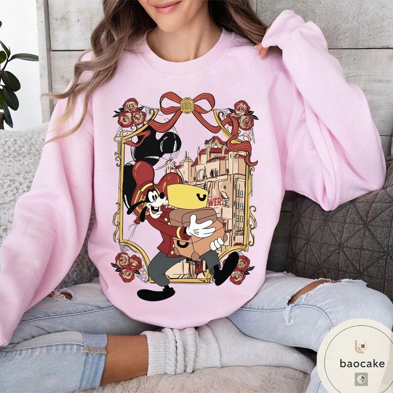 May include: A light pink sweatshirt featuring a cartoon character carrying luggage in front of a building. The character is wearing a red jacket and hat. The design is framed with roses and a bow.