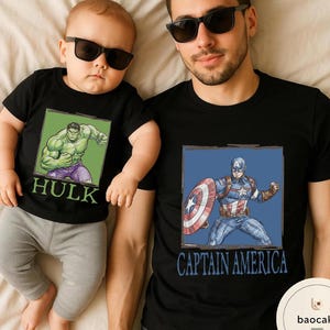 May include: Black t-shirts with superhero graphics. One shirt features the Hulk, the other Captain America. The Hulk shirt is worn by a baby, and the Captain America shirt is worn by an adult. Both shirts have a square graphic.