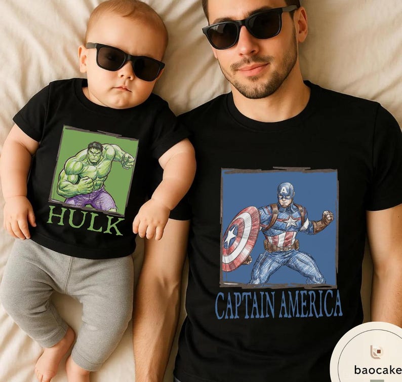 May include: Black t-shirts with superhero graphics. One shirt features the Hulk, the other Captain America. The Hulk shirt is worn by a baby, and the Captain America shirt is worn by an adult. Both shirts have a square graphic.