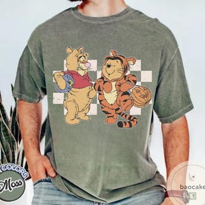 May include: A moss green t-shirt featuring a cartoon illustration of two characters dressed in Halloween costumes. One character is dressed as a bear and the other as a tiger. The bear is holding a blue bag, and the tiger is holding a jack-o'-lantern.