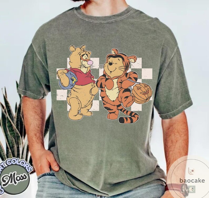 May include: A moss green t-shirt featuring a cartoon illustration of two characters dressed in Halloween costumes. One character is dressed as a bear and the other as a tiger. The bear is holding a blue bag, and the tiger is holding a jack-o'-lantern.