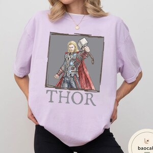 May include: A light purple t-shirt featuring a graphic of Thor holding his hammer, Mjolnir, within a gray rectangle. The word "THOR" is printed in gray below the graphic. The shirt has short sleeves and a crew neck.
