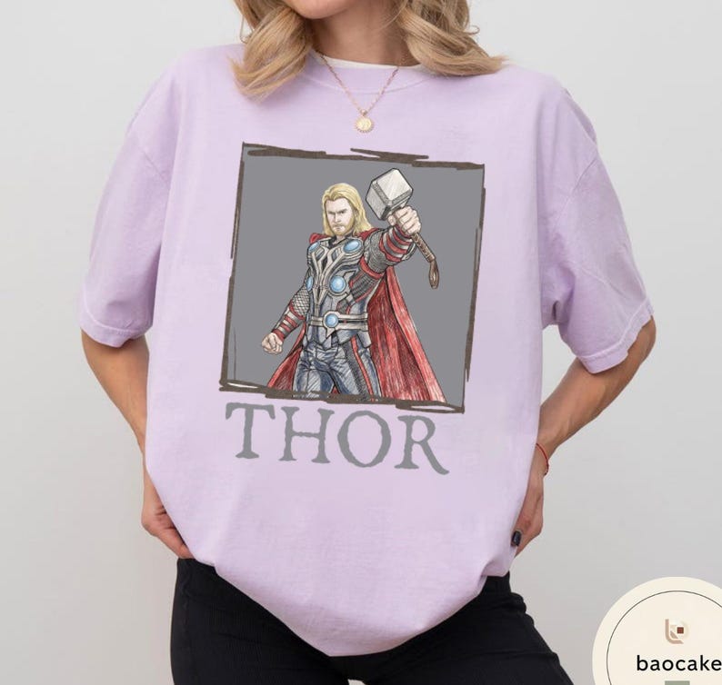 May include: A light purple t-shirt featuring a graphic of Thor holding his hammer, Mjolnir, within a gray rectangle. The word "THOR" is printed in gray below the graphic. The shirt has short sleeves and a crew neck.