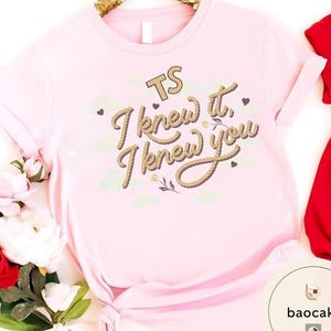 K&ouml;nnte beinhalten: Ein hellrosa T-Shirt mit dem Text TS I knew it, I knew you in einer braunen Schriftart im Seil-Stil. Das Design enth&auml;lt kleine braune Herzen und hellgr&uuml;ne Wolkenformen. Das T-Shirt hat kurze &Auml;rmel und einen geknoteten Saum am unteren Rand.