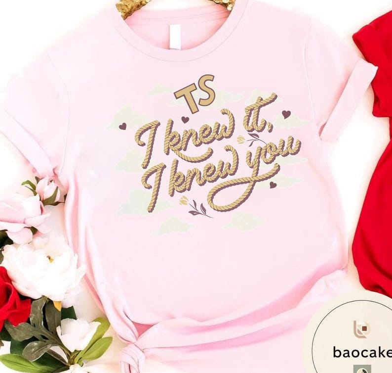 K&ouml;nnte beinhalten: Ein hellrosa T-Shirt mit dem Text TS I knew it, I knew you in einer braunen Schriftart im Seil-Stil. Das Design enth&auml;lt kleine braune Herzen und hellgr&uuml;ne Wolkenformen. Das T-Shirt hat kurze &Auml;rmel und einen geknoteten Saum am unteren Rand.